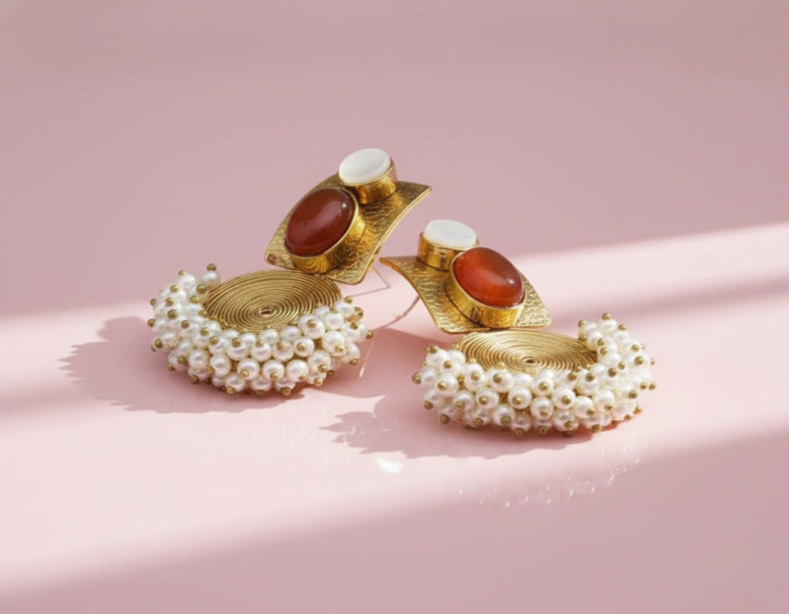 Pair of gold earrings with pearl and red stone accents on a pink background